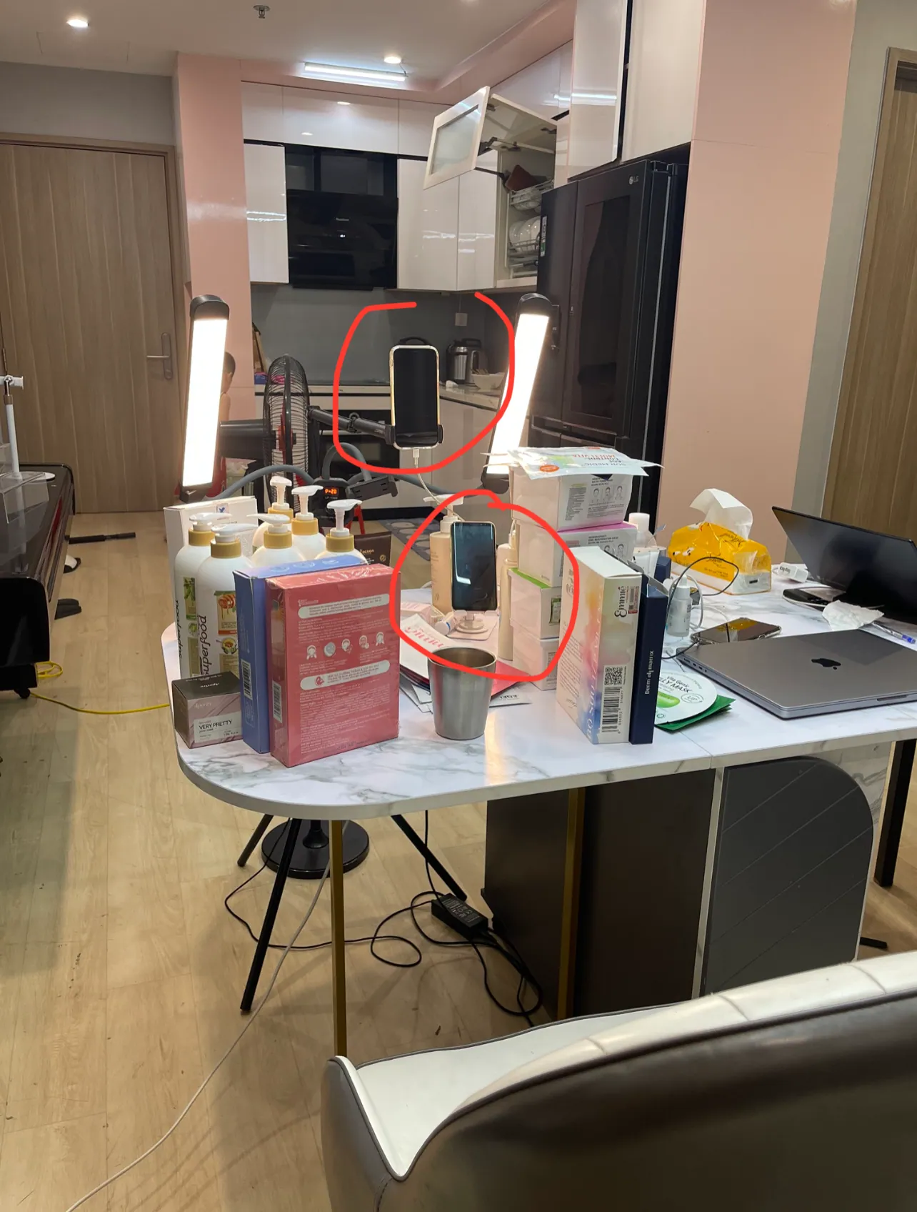 Góc phòng bếp được dựng thành chỗ livestream: bàn đá phủ kín chai lọ mỹ phẩm và hộp giấy, hai đèn thanh dựng hai bên, một điện thoại kẹp trên giá và một điện thoại thứ hai đặt trên bàn.