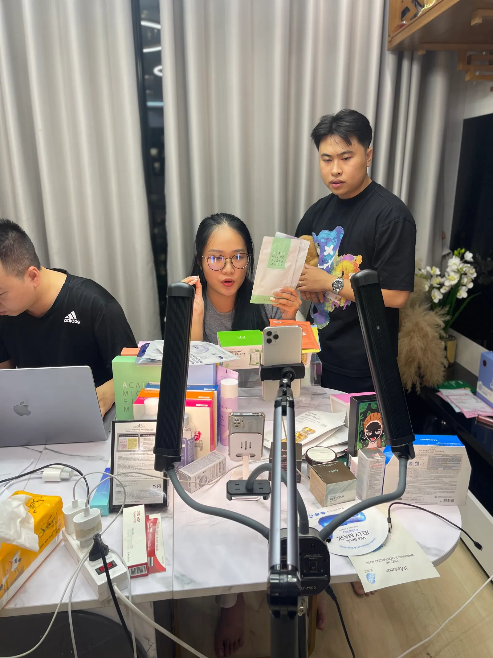 Ba người ngồi và đứng quanh một chiếc bàn phủ kín sản phẩm, có giá kẹp điện thoại và đèn livestream, hai laptop mở sẵn.
