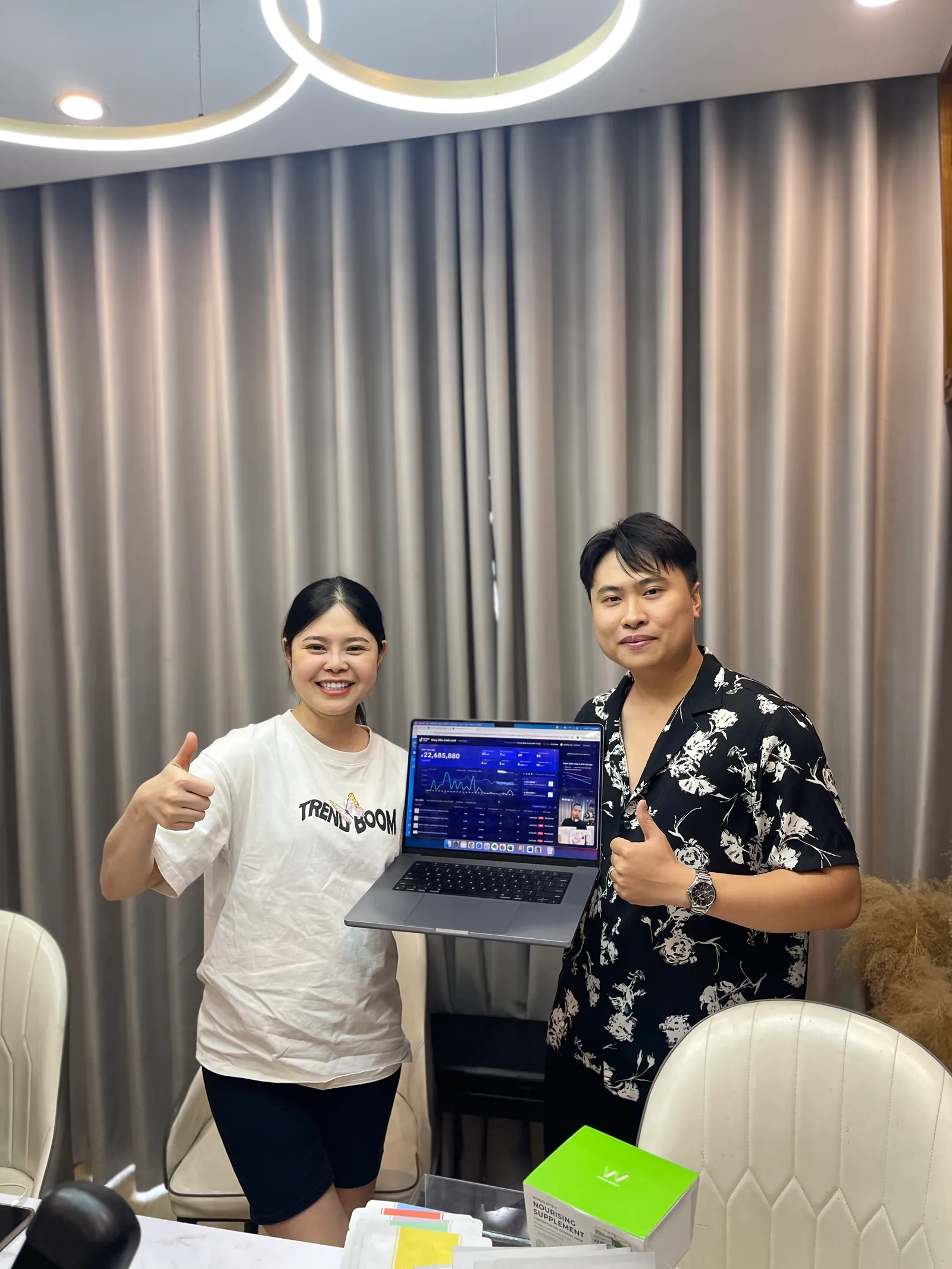 Hai người đứng trong phòng livestream cùng giơ ngón tay cái, cầm một chiếc laptop đang mở bảng theo dõi doanh thu của phiên bán.